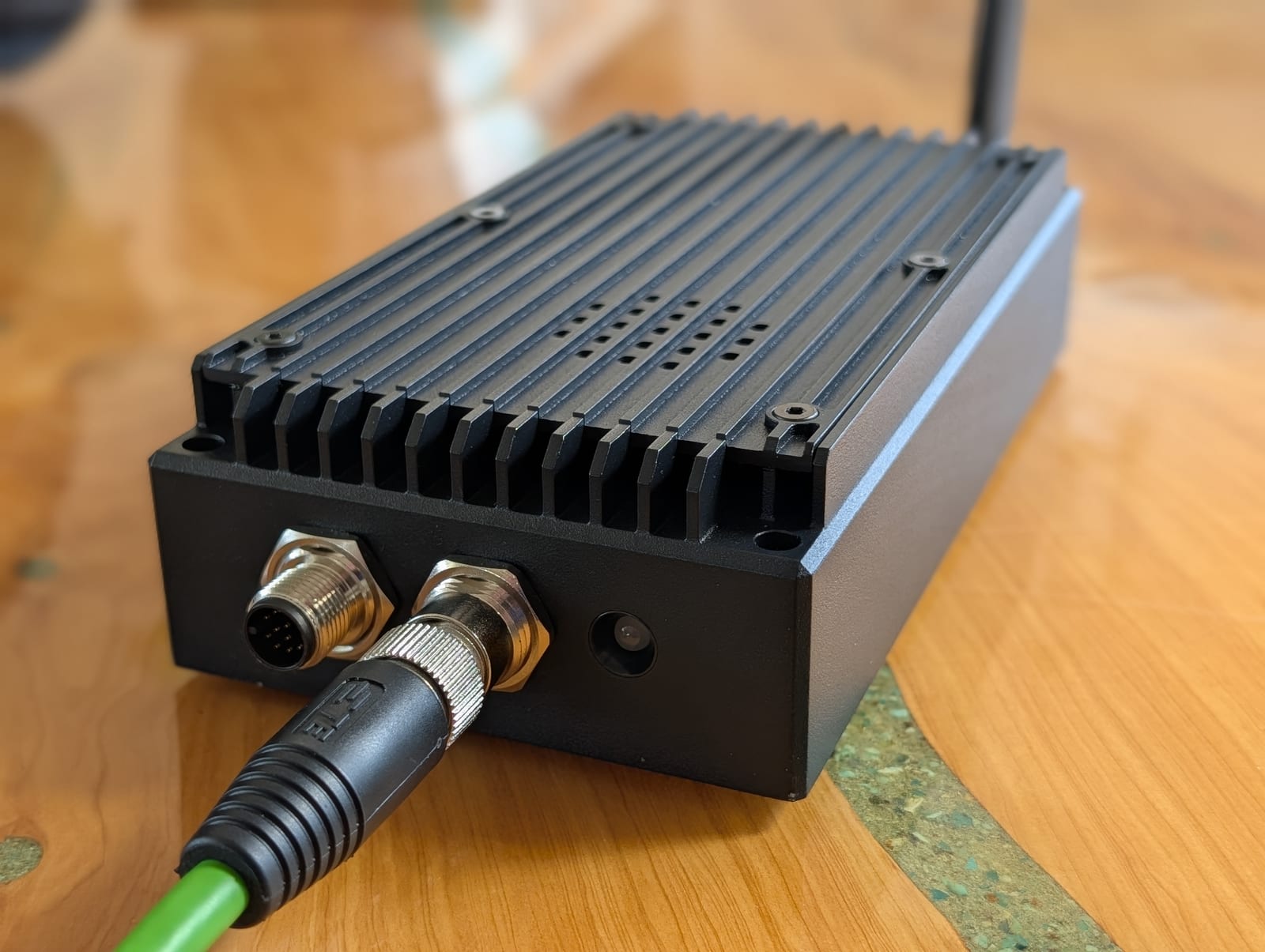 Front three-quarter view of the COYOTE unit: a black anodized ruggedized enclosure with machined heat-sink fins, sealed M12 connectors with an Ethernet cable attached, and a whip antenna.